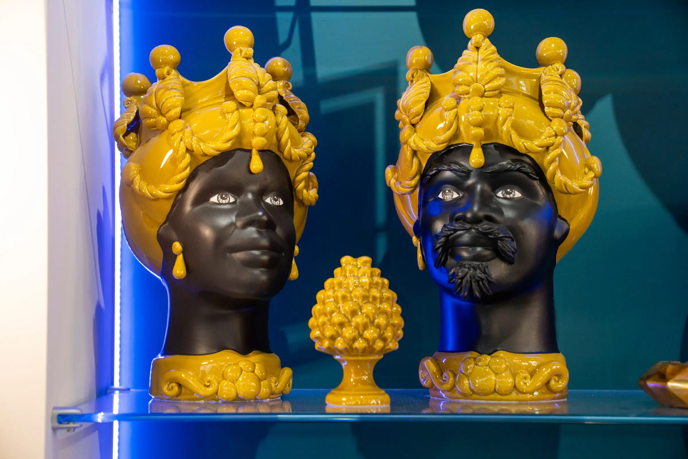 Due teste di moro in ceramica smaltata, nere con corone gialle, e una pigna gialla su una mensola illuminata di blu.